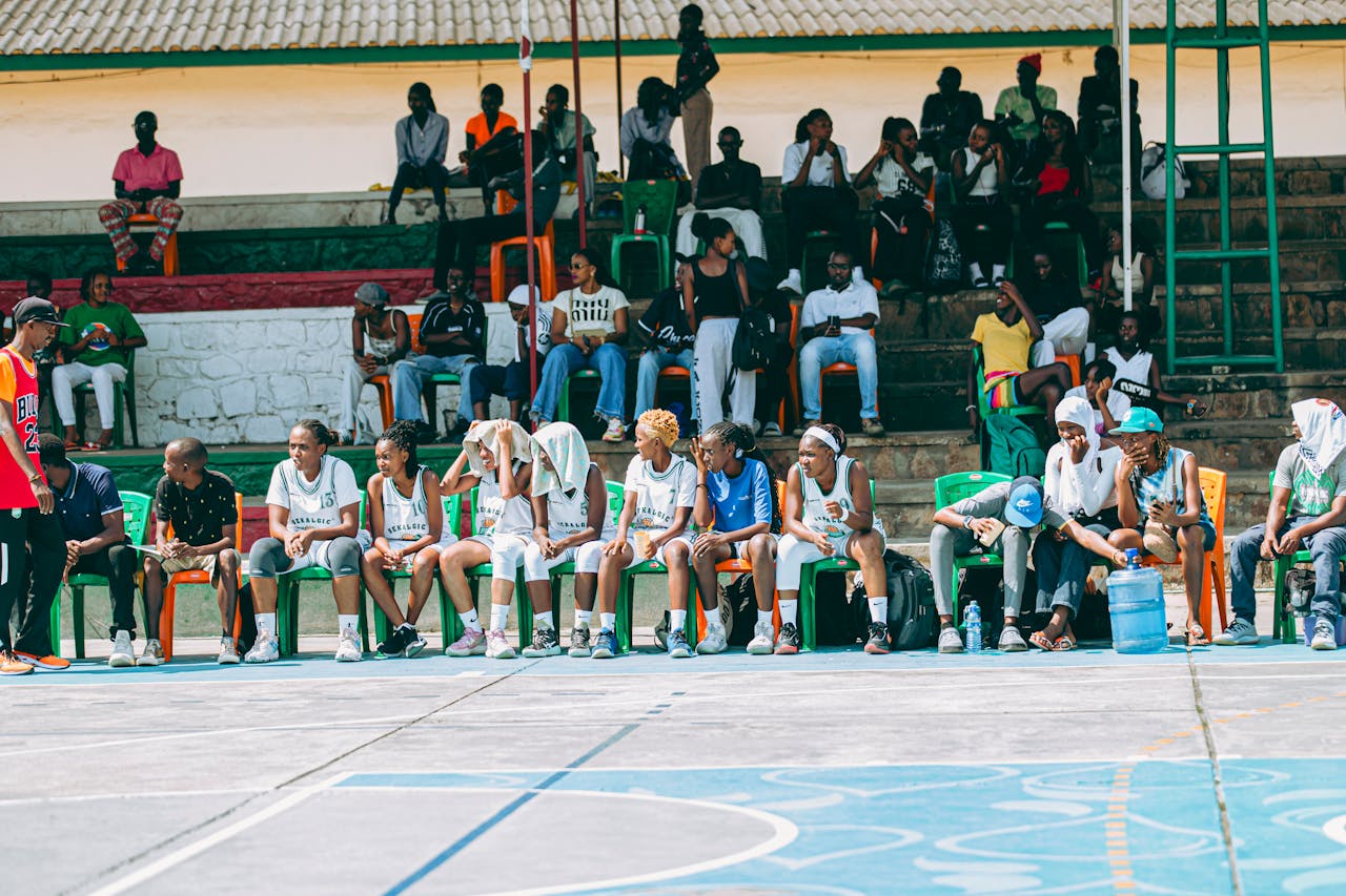brand-01 Team sitting on sidelines at outdoor basketball game with spectators in background.